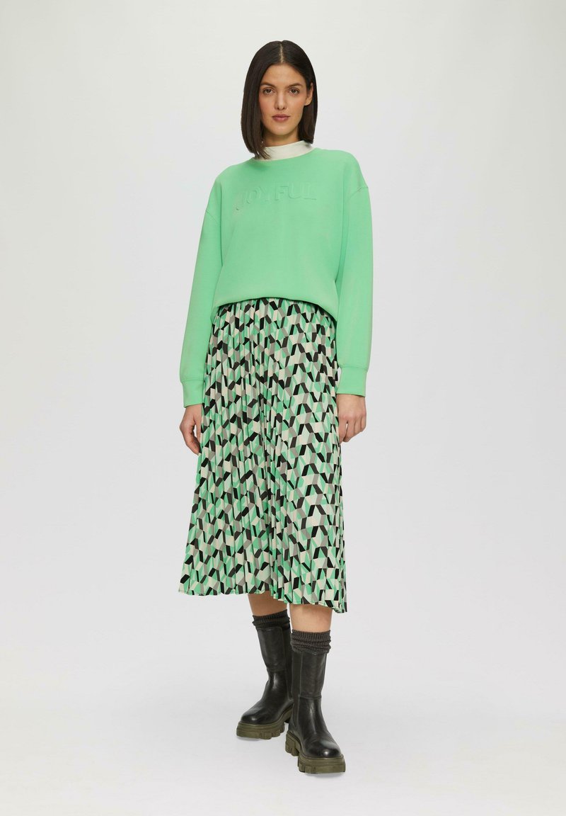 Sweatshirt vert menthe avec le mot "JOYFUL" en relief, associé à une jupe midi à motifs géométriques dans des tons vert, noir et crème. Bottes noires jusqu'aux genoux.