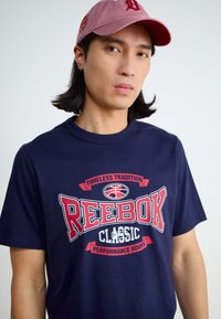 Marineblå bomulls t-skjorte med en dristig Reebok-grafikk i rødt og hvitt, rund halsutskjæring, avslappet passform, brukt sammen med en rosa baseball caps.