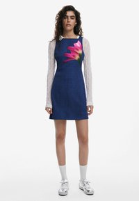 Rochie din denim albastru cu un model floral pe față, cămașă albă cu mâneci lungi dedesubt și adidași argintii. Textură fină și design casual.