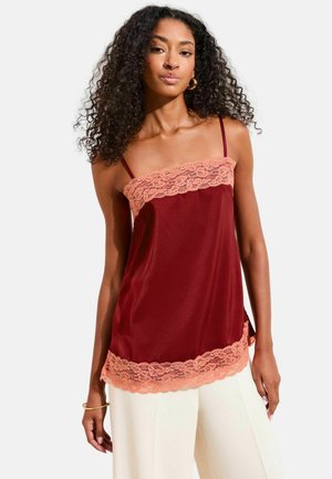 Kvinde iført en ærmeløs bordeaux satin top med ferskenfarvet blondekant, kombineret med hvide bukser med vide ben, stående foran en ensfarvet baggrund.