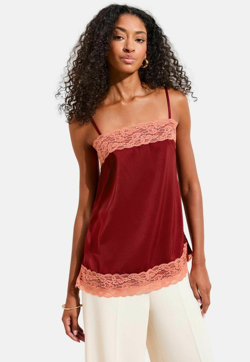 Femeie purtând un top fără mâneci din satin burgundy cu dantelă piersică, asortat cu pantaloni albi cu picioare largi, stând în fața unui fundal simplu.