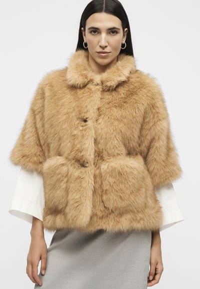 Femme portant une veste en fausse fourrure beige à manches courtes sur des manches longues blanches et une jupe grise, regardant devant avec une expression neutre.