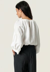 Hvid, tekstureret bluse med høj hals og rykke med nøglehul-detail, sammen med grå stribede bukser. Løs pasform og indsamlede ærmer.