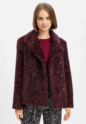 Young woman wearing a burgundy textured double-breasted coat, burgundy top, and black patterned pants, standing against a plain white background.