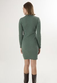 Abito maglione verde a treccia con silhouette aderente, maniche lunghe e polsini a coste, che arriva a metà coscia.