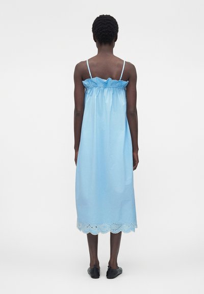 Άτομο με κοντά σγουρά μαλλιά, που φοράει αμάνικο γαλάζιο φόρεμα μήκους μέσης με διακοσμητική δαντέλα στο τελείωμα και μαύρα επίπεδα παπούτσια, στέκεται με την πλάτη προς τα εμπρός.