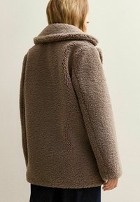 En kvinna med ljusbrunt hår har på sig en tjock, brun teddyjacka med bred krage, sedd bakifrån mot en enkel ljus vägg.