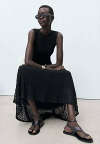 Robe maxi en dentelle noire avec un ourlet superposé, portée avec des sandales noires élégantes. Le mannequin porte des lunettes de soleil noires angulaires et des bracelets en or.