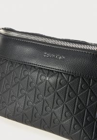 Clutch bag nera in pelle con motivi a triangolo e cerchio in rilievo, chiusura con zip argentata e logo "Calvin Klein" in argento.