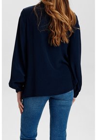 Blouse bleu marine avec de longues manches volumineuses, texture lisse et silhouette décontractée, associée à un jean bleu ajusté.