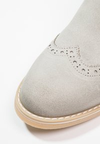 Gros plan sur l'avant d'une chaussure en daim gris clair avec des détails de perforations brogue et une semelle beige clair avec des coutures blanches visibles.
