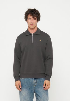 Homme aux cheveux bouclés portant un sweat zippé gris foncé avec un petit logo jaune et un jean bleu clair, debout sur un fond blanc uni.