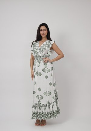Kobieta stojąca z jedną ręką na biodrze, ubrana w białą suknię maxi w zielone kwiatowe wzory z falbankami na rękawach, na jednolitym tle.
