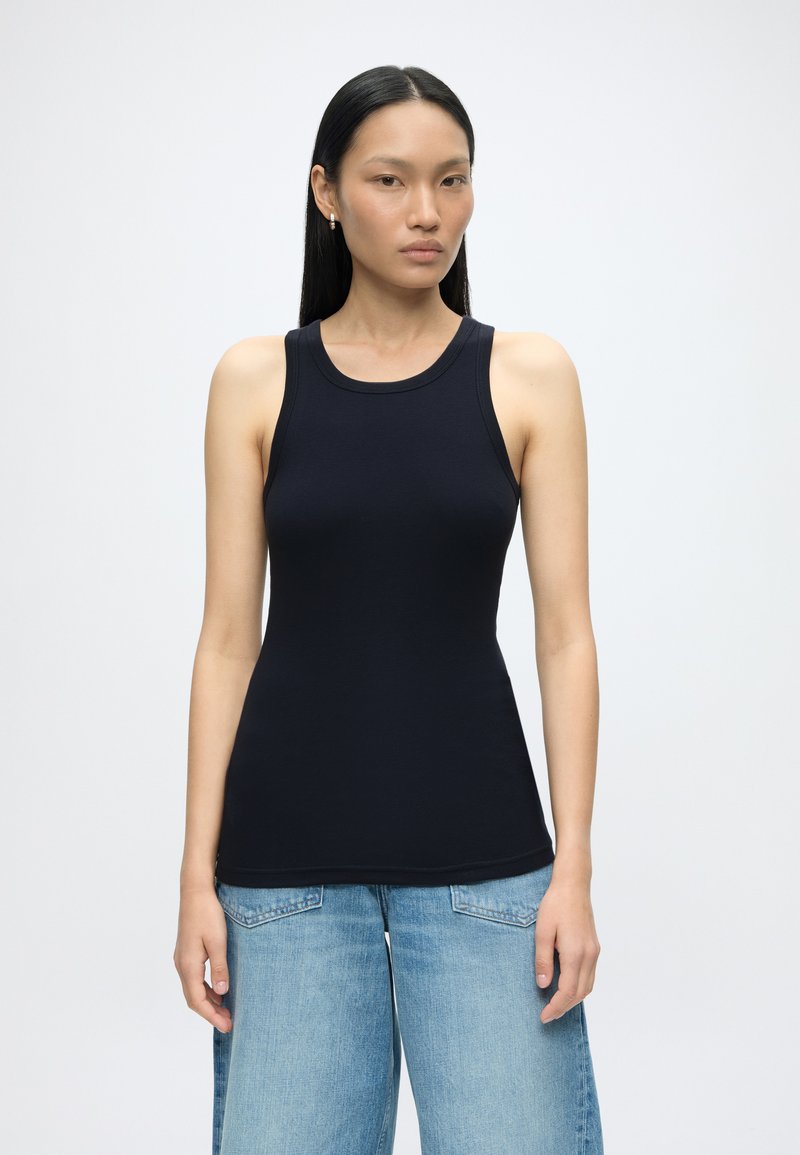 Vrouw met lang zwart haar, gekleed in een aansluitend zwart mouwloos topje en lichtblauwe wijde jeans, staand tegen een effen witte achtergrond.