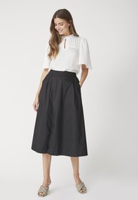 Blusa bianca con texture e scollo a chiave, abbinata a una gonna nera di lunghezza media con pieghe e tasche, indossata con sandali grigi.