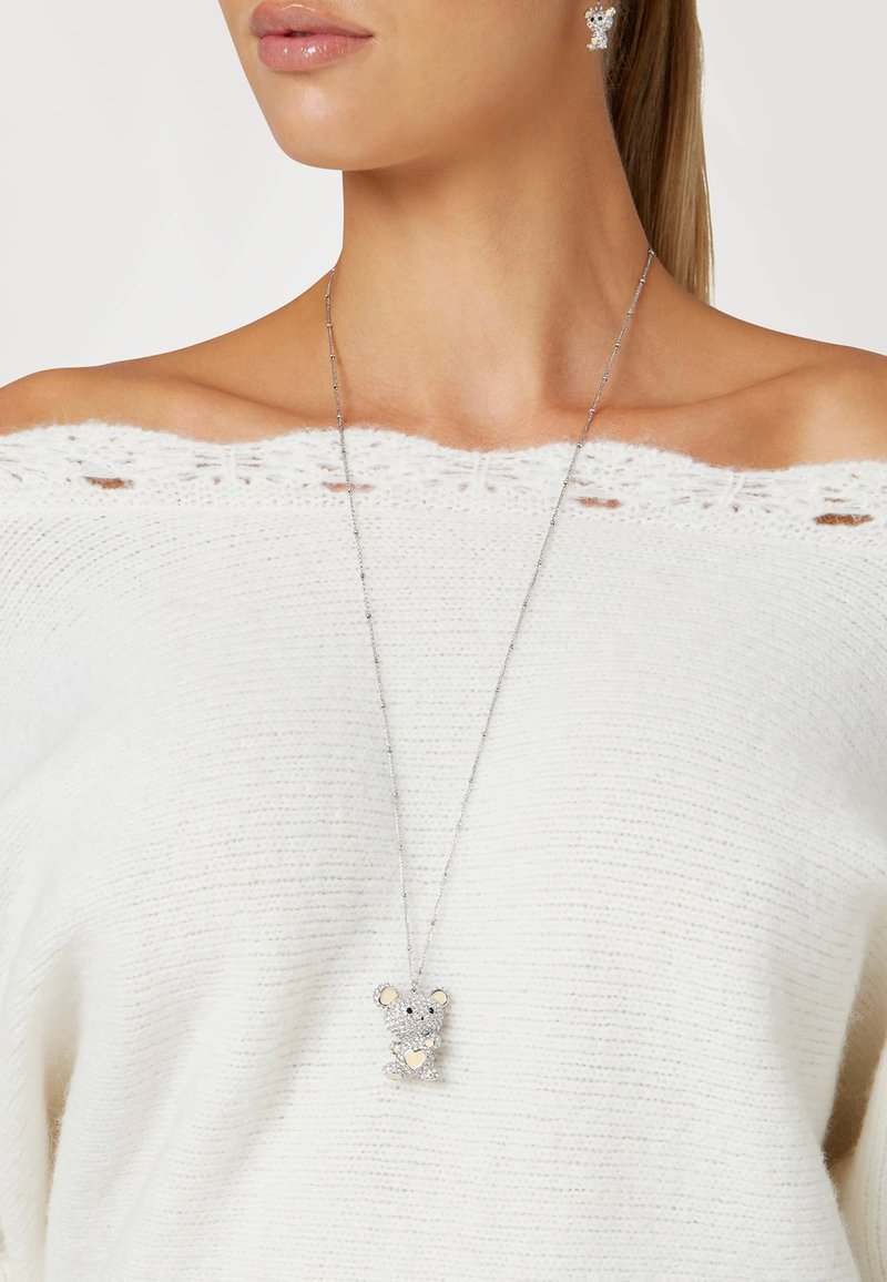 Zilveren ketting met een berenhanger, voorzien van heldere rhinestones en gouden accenten, gedragen over een witte off-shoulder gebreide trui.