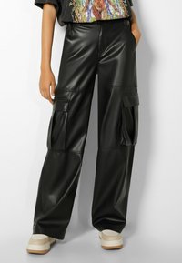 Czarne spodnie cargo z ekoskóry o szerokich nogawkach, z dwoma przednimi kieszeniami i gładką teksturą, połączone z jasnymi sneakerami.