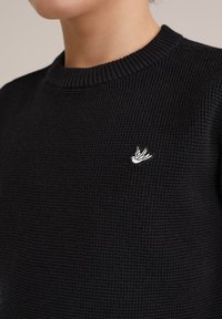Črna pletena jopica z rebrastim okroglim izrezom, z majhnim belim vezenim logotipom ptice na levi strani. Teksturirana tkanina.