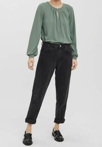 Blouse verte à manches longues avec un col en forme de goutte d'eau, associée à un jean en denim noir taille haute, retroussé aux chevilles, et des mocassins noirs.