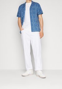 Modrá košile s krátkým rukávem a geometrickým vzorem, v kombinaci s bílými kalhotami a bílými teniskami. Hladká textura a neformální design.