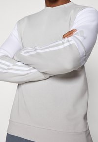 Sweat-shirt gris clair avec manches longues blanches, comportant trois bandes blanches le long des bras et un col rond ; texture de tissu douce.