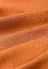 Textil in einem kräftigen Orangeton mit einer glatten Textur, sanft mit weichen Falten drapiert. Leicht strukturierte Oberfläche mit subtilen Variationen.