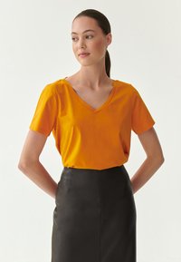 T-shirt s výstřihem do V a krátkým rukávem v jasně oranžové bavlně, doplněný černou koženou tužkovou sukní, hladká textura a přiléhavý střih.