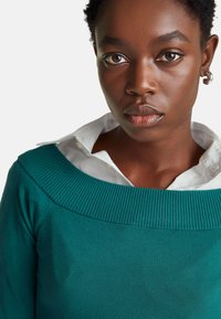 Tyrkysový svetr s žebrovaným výstřihem, nošený přes bílou košilí s límečkem. Vyznačuje se hladkou texturou a přiléhavým střihem, který zvýrazňuje čistou siluetu.