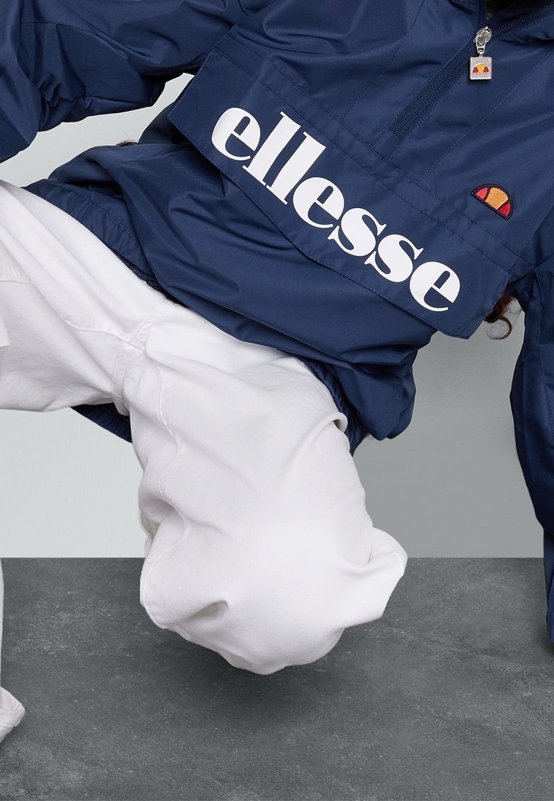 Personne portant une veste ellesse bleu marine et un pantalon blanc dans une pose accroupie dynamique sur une surface grise.