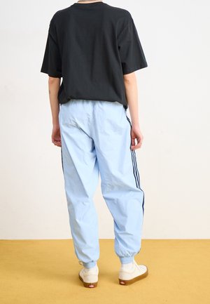Person trägt ein schwarzes oversized T-Shirt, hellblaue Jogginghosen mit dunklen Streifen und weiße Turnschuhe, steht auf einem gelben Boden vor einer weißen Wand.