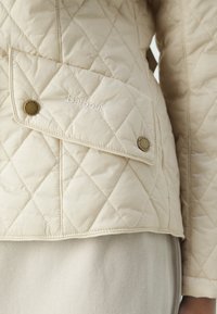Casaco bege acolchoado com um bolso com aba, logotipo em relevo e ferragens gravadas, destacando uma textura suave e um padrão de losangos.