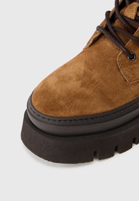 Bota de tobillo de ante marrón con suela negra gruesa. Presenta punta redondeada, ojales de metal y cordones oscuros, destacando el material texturizado.