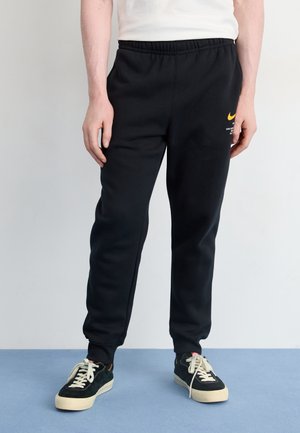 Personne portant un pantalon de survêtement noir Nike avec un logo jaune, un t-shirt blanc et des baskets noires avec des lacets blancs, debout sur un sol bleu.