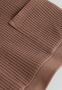 Brauner, strukturierter Stoff mit Waffelmuster, der einen glatten Akzentbereich aufweist, hervorgehoben durch eine gefaltete Kante und einen kontrastierenden Besatz.