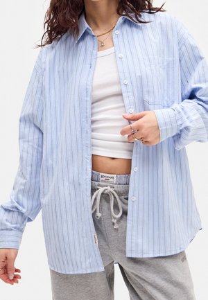 Person iført en lyseblå stribet skjorte over en hvid tanktop og grå joggebukser med snøre, der viser en afslappet, casual stil.