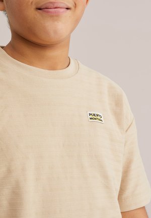 Közelkép egy személyről, aki bézs színű, texturált pólót visel, amelyen a mellrészen egy kis "Puerto Montana" feliratú címke látható, egyszínű háttér előtt.