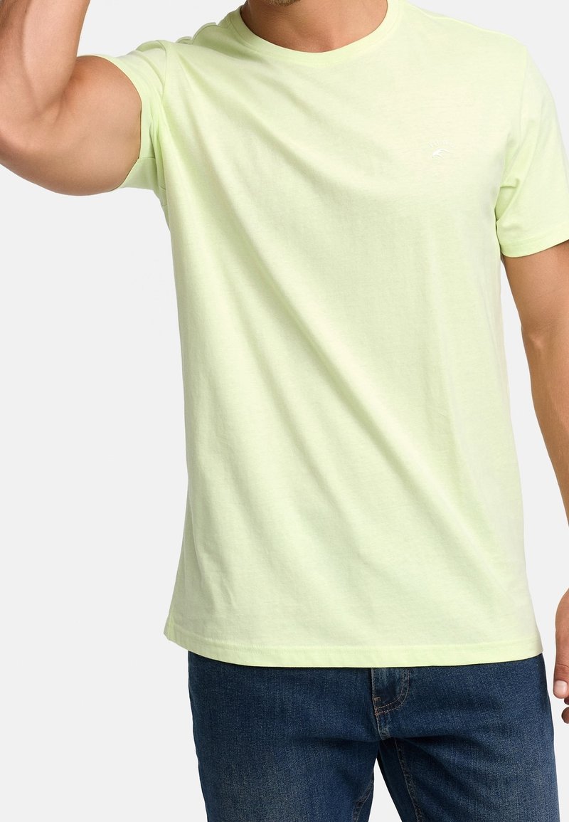 Homme portant un T-shirt à manches courtes jaune clair et un jean bleu, levant un bras devant un fond uni.
