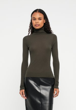 Vrouw met lang krullend haar draagt een aansluitende olijfgroene coltrui en een zwarte leren rok, staand tegen een effen lichte achtergrond.