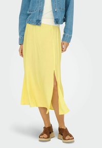 Falda amarilla, ligera y de longitud media con una abertura lateral. Llevada con una chaqueta de mezclilla y combinada con sandalias marrones de tiras sobre una plataforma texturizada.