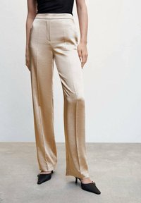 Pantalones rectos de satén beige con una textura suave, pliegues delanteros y una cintura alta, combinados con tacones negros puntiagudos.