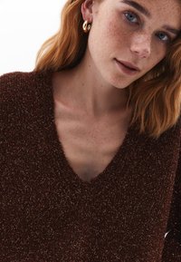 Maglione a maglia marrone con texture e scollatura a V. La modella indossa orecchini a cerchio dorati, mostrando lentiggini e una pelle chiara.
