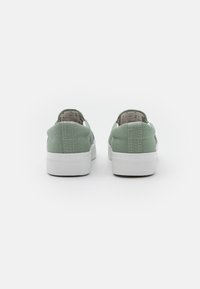 Zapatillas verdes de lona tipo slip-on con suela de goma blanca, que cuentan con detalles de costura y una parte trasera texturizada para un mejor agarre.
