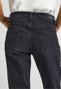 Jeans de mezclilla negros con cintura de altura media, cinco bolsillos y una etiqueta de logo simple en la parte posterior. La tela tiene una textura suave con un desvanecido mínimo.