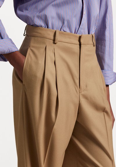 Beige plisserade byxor i mjukt tyg med hög midja och sidofickor, kombinerade med en blå och röd randig skjorta.