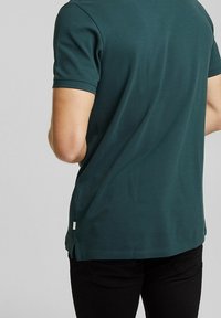 Donkergroene poloshirt met korte mouwen, gestructureerde stof, zijsplitten aan de zoom en een klein wit label linksonder.