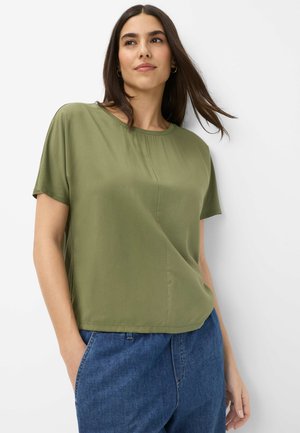 Woman with long dark hair wearing olive green short-sleeve blouse and blue jeans, standing against a white background.