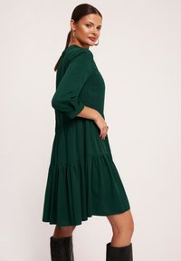 Zielona, sukienka do kolan z zwiewnego materiału z marszczonymi warstwami, rękawami 3/4 i okrągłym dekoltem. Połączona z czarnymi kozakami sięgającymi do kolan.