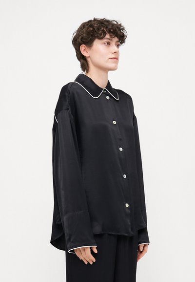Black satin shirt with a rounded collar, white piping, long sleeves, and button front. Smooth texture, relaxed fit, and side slits.