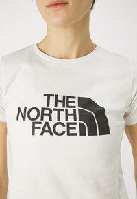 Biała koszulka z bawełny z dużym czarnym logo z napisem "THE NORTH FACE" w pogrubionej, wielkiej literze. Krótkie rękawy, okrągły dekolt.