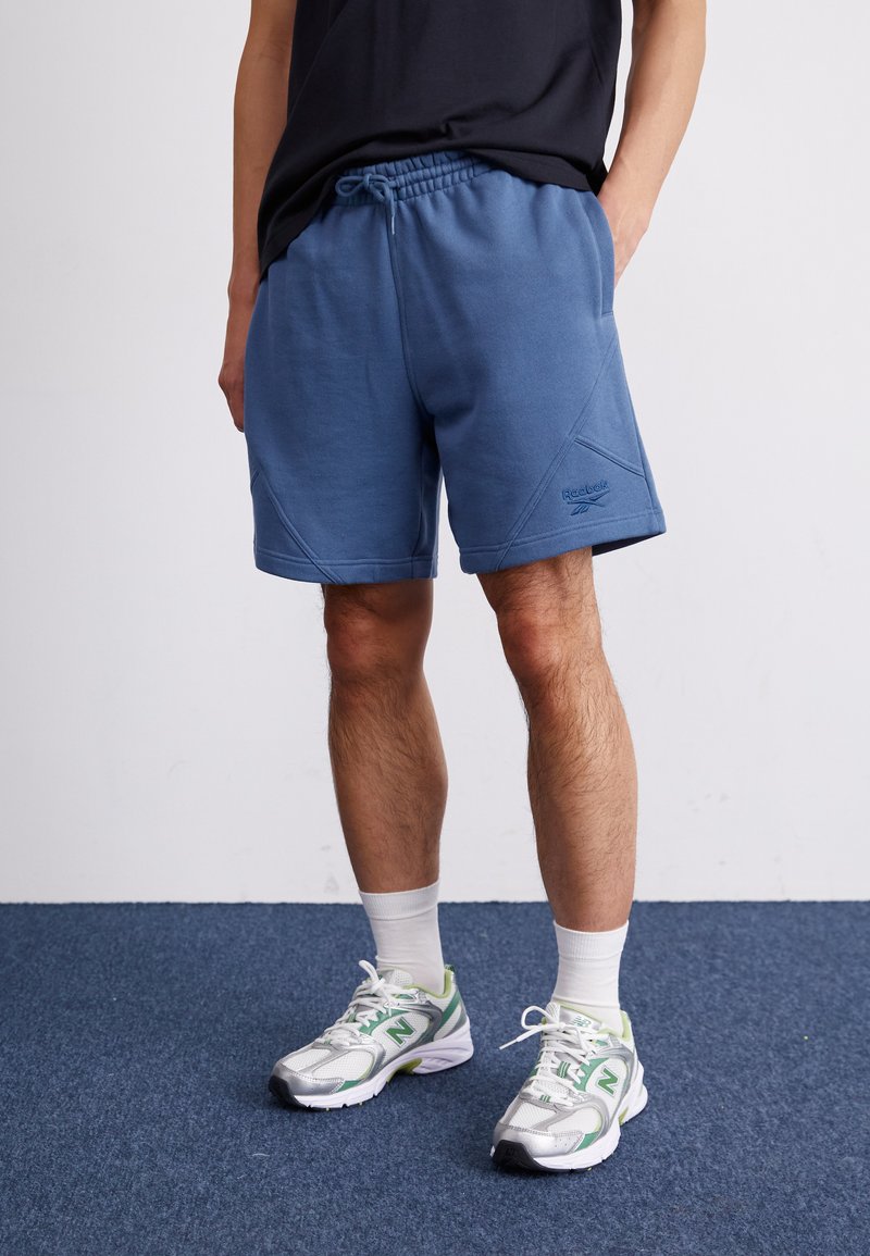 Shorts de sport bleu avec taille à cordon, en tissu texturé, coutures diagonales et un petit logo sur le côté gauche. Assortis avec des chaussettes blanches et des baskets.